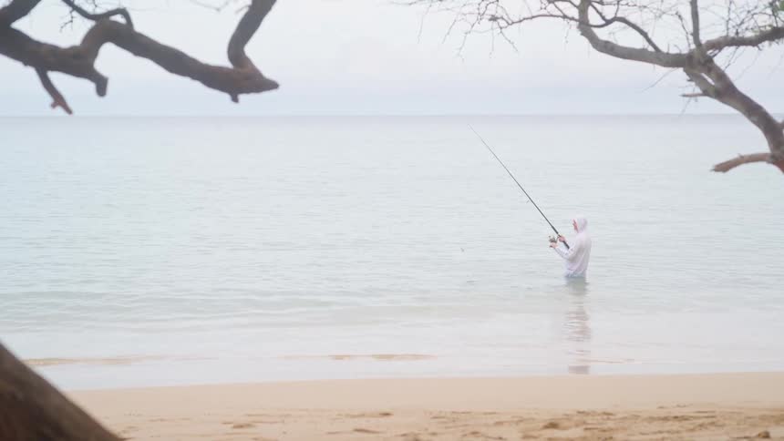 A fisherman in a white hooded shirt stands in the shallow ocean water, patiently fishing on a calm, overcast day