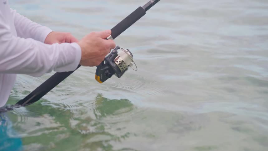 A close-up shot of a fisherman reeling in his line while standing in calm, shallow water