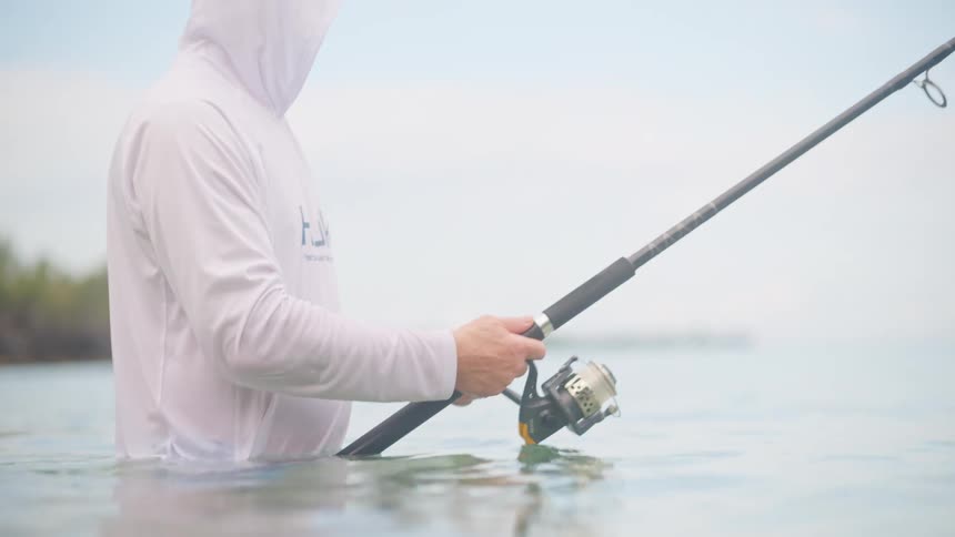 A fisherman wearing a white hooded shirt stands in calm ocean water, reeling in his fishing line