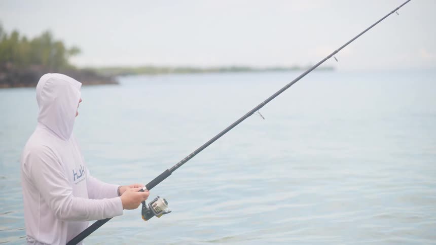 A man in a white hooded shirt stands in the calm ocean water, patiently reeling in his fishing line on an overcast day