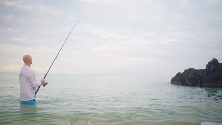 A man patiently waits for a catch while surf fishing in the calm ocean shallows on an overcast day