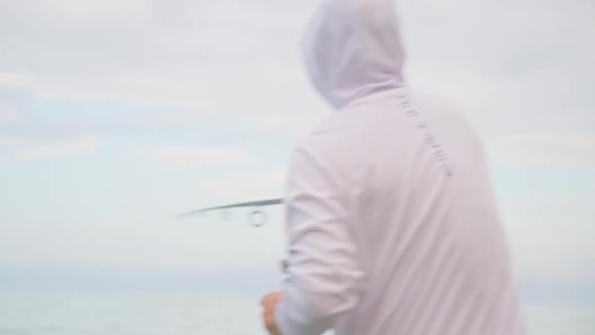 A man in a white fishing hoodie casts his line into the calm ocean in slow motion