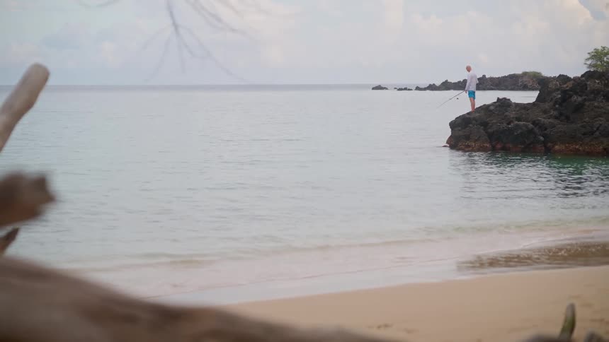 A man enjoys a peaceful day of fishing from the rocks on a tranquil tropical beach