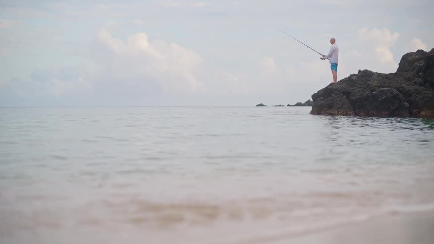A man enjoys a peaceful moment of solitude, fishing from the rocks as ocean waves gently break on the beach