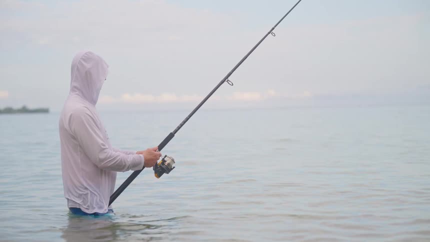 A man in a white hooded shirt patiently fishes while standing in the calm, shallow ocean on an overcast day