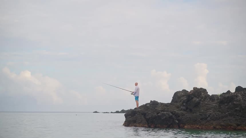 A lone fisherman stands on the rocks by the calm ocean on an overcast day