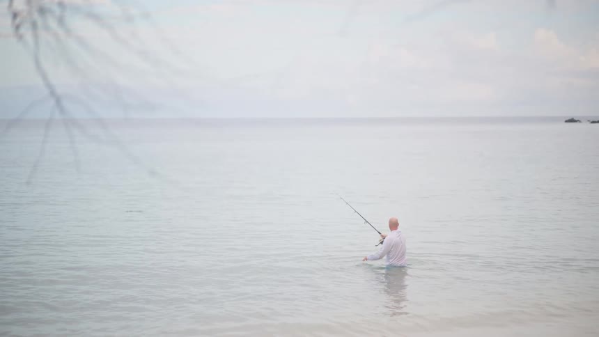 A solitary fisherman in a white shirt stands waist-deep in the calm, pale ocean, casting his line on an overcast day