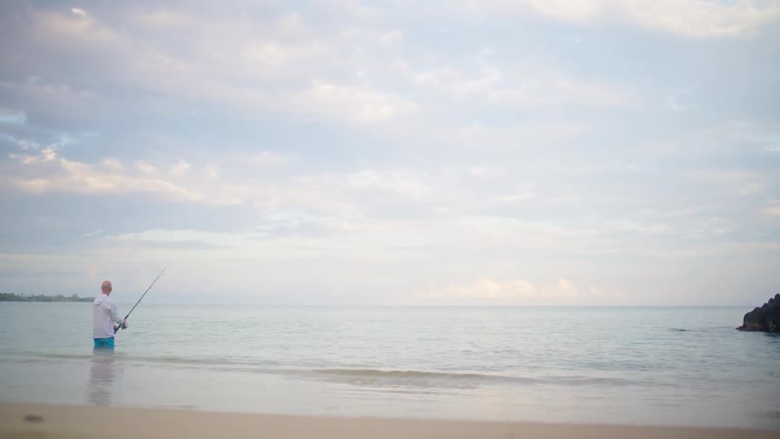 A lone fisherman stands in the calm ocean shallows, fishing from a sandy beach on an overcast day