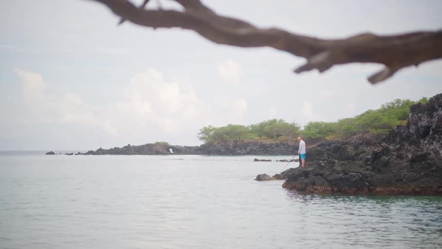 A lone fisherman stands on a rocky volcanic shoreline, patiently fishing in the calm ocean on a quiet, overcast day