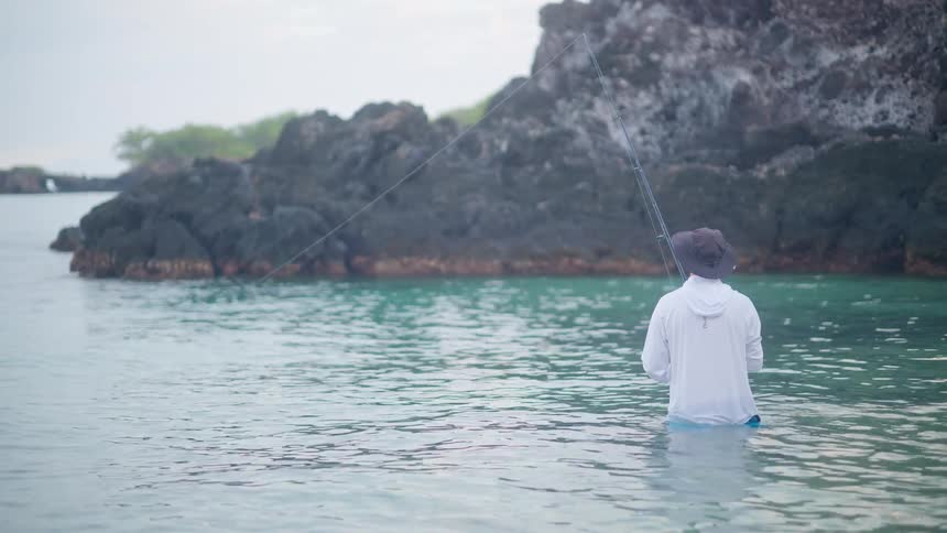 A fisherman stands in the calm, clear ocean water, patiently holding a fishing rod near a rocky shoreline