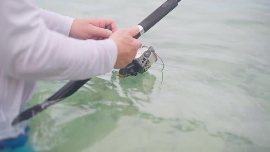 Close-up of a fisherman's hands reeling in a fishing rod while standing in clear, shallow ocean water