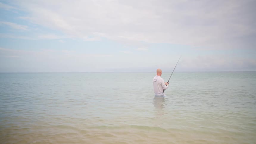 A solitary fisherman casts his line into the calm, endless ocean from the shallow waters of a serene beach