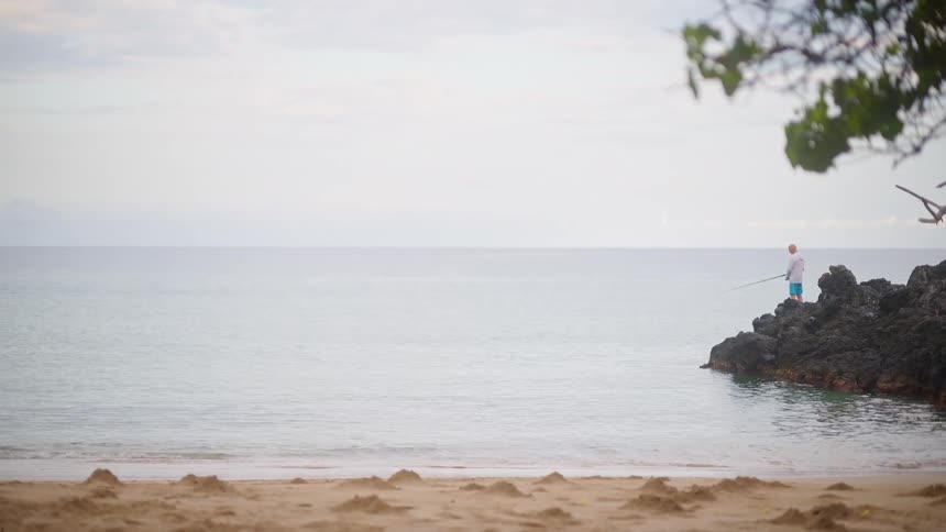 A lone fisherman patiently casts his line into the calm ocean from a rocky point on a sandy beach