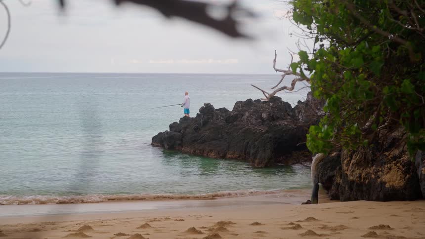 A solitary fisherman stands on a rocky outcrop on a tropical beach, casting his line into the calm ocean