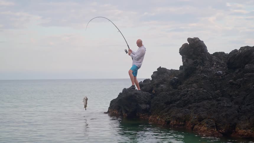 A fisherman standing on a rocky shore reels in a fish from the ocean in dramatic slow motion