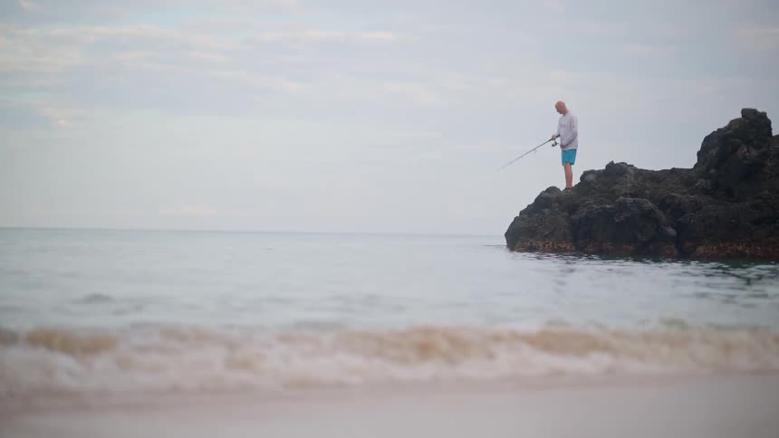 A lone fisherman stands on a rocky outcrop in the calm ocean, patiently waiting for a catch as waves gently roll onto the shore