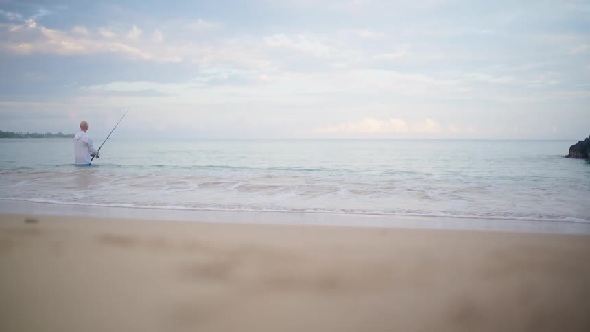 A lone fisherman stands in the shallow ocean waters, fishing from the shore as gentle waves roll in on a tranquil day