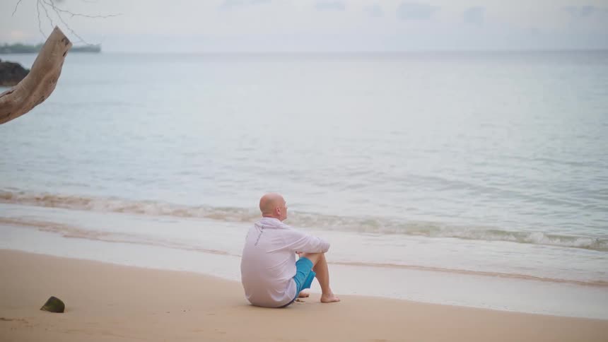 A lone man sits on a sandy beach, peacefully watching the ocean waves roll onto the shore