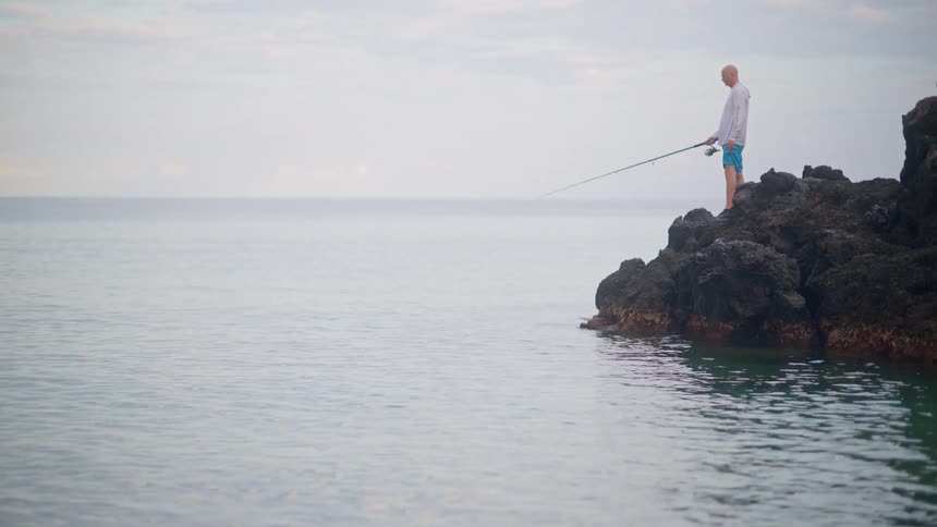 A lone fisherman stands patiently on a rocky coast, casting his line into the calm, quiet ocean