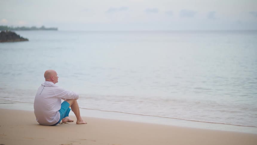 A contemplative man sits alone on a quiet sandy beach, watching the calm ocean waves
