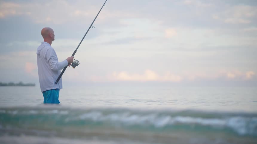 A lone fisherman stands in the tranquil ocean at dawn, patiently waiting with his fishing rod as gentle waves lap the shore