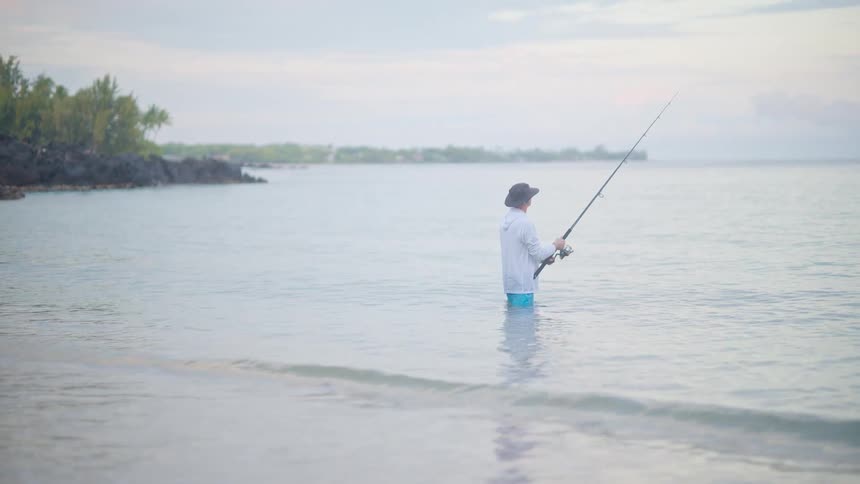 A lone fisherman stands in the shallow, calm ocean water, patiently fishing on a tranquil, overcast day