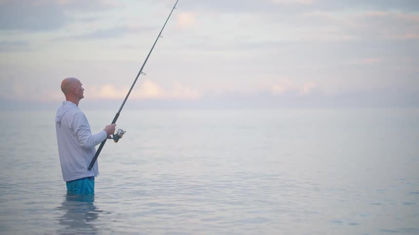 A solitary fisherman patiently waits for a bite while standing waist-deep in the tranquil ocean at sunrise
