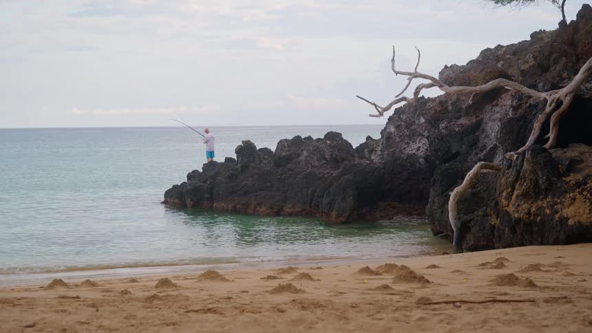 A lone fisherman stands on a rocky outcrop of a sandy beach, fishing in the calm ocean water