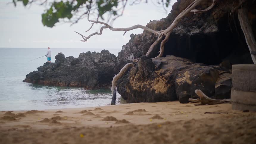 A lone fisherman casts his line into the calm ocean while standing on a rocky outcrop of a sandy beach