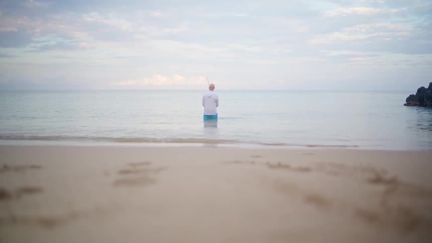 A lone fisherman stands in the tranquil ocean shallows as gentle waves lap the sandy shore