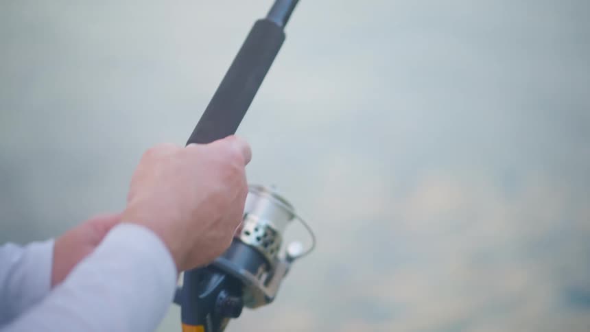 A slow-motion close-up focuses on a fisherman's hands as he reels in his line, panning up the rod to the tip