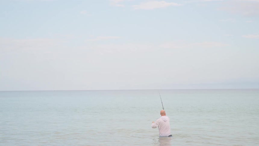 A lone fisherman casts his line while standing waist-deep in the tranquil ocean on a calm day
