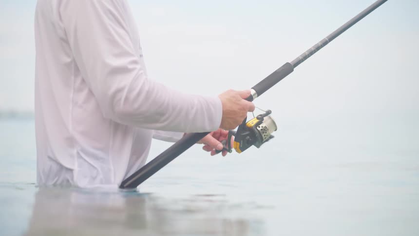 A fisherman patiently reels in his line while standing in the shallow, calm sea on an overcast day