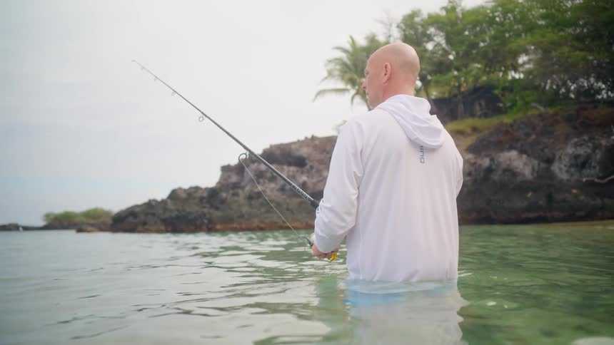 A fisherman stands patiently waist-deep in the serene, clear water of a tropical bay on an overcast day