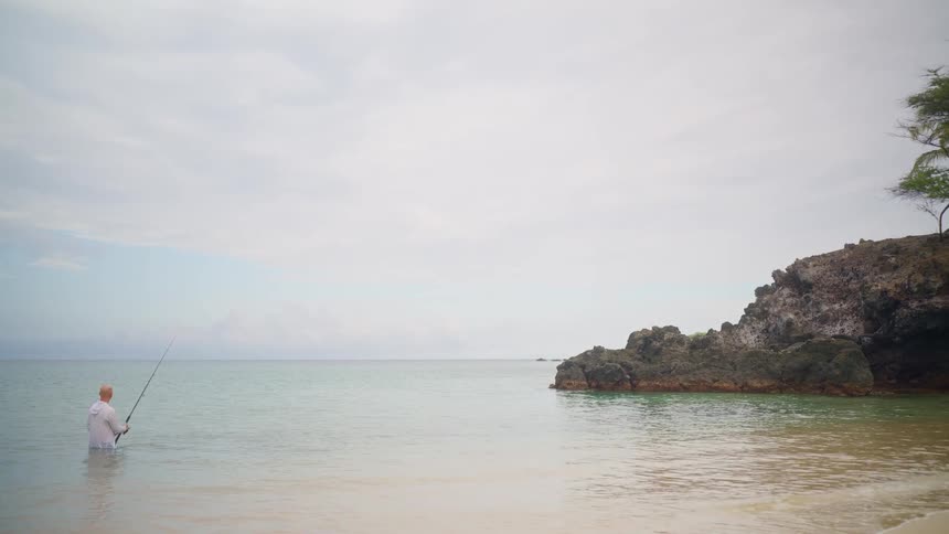 A lone fisherman stands in the turquoise ocean, casting his line from a peaceful sandy beach with a rocky shoreline