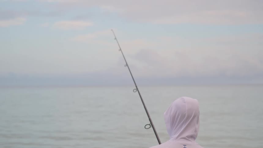 A lone fisherman in a white hoodie patiently waits for a bite while looking out over the tranquil sea under a soft, hazy sky