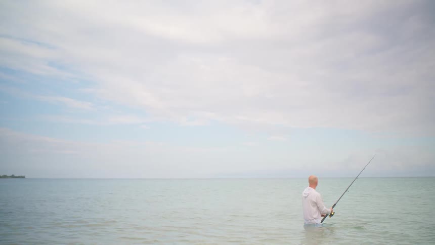 A lone fisherman patiently waits for a catch while standing in the calm, tranquil sea on an overcast day