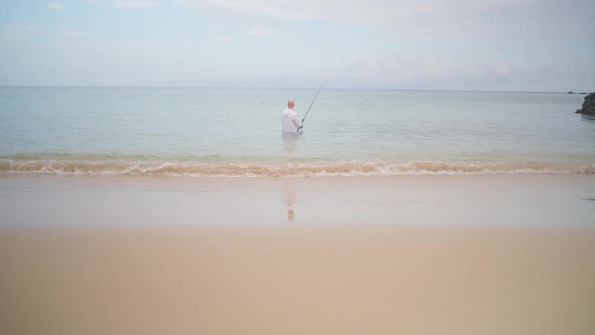 A man stands peacefully in the calm ocean water fishing as gentle waves roll onto the sandy shore