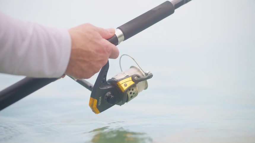 A close-up slow motion shot shows a fisherman's hand holding a fishing rod and reel in the calm, clear water