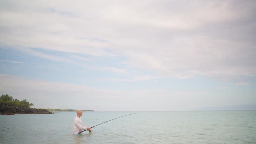 A man stands waist-deep in the calm ocean water and casts his fishing rod on a beautiful day