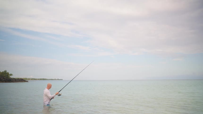 A bald man in a white shirt stands waist-deep in the calm ocean water, reeling in his fishing line