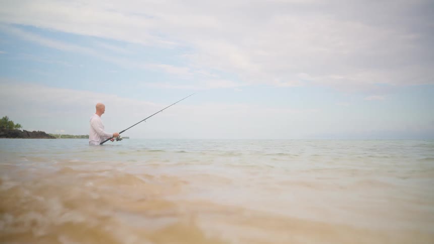 A man patiently fishes in the shallows of the ocean as gentle waves lap the shore in slow motion