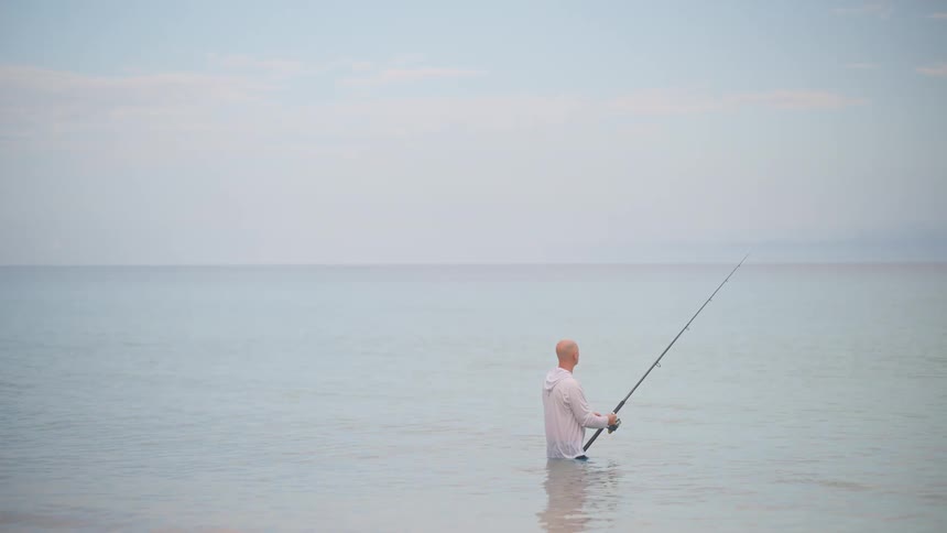 A man stands waist-deep in the calm ocean, patiently fishing with a rod under a quiet, overcast sky