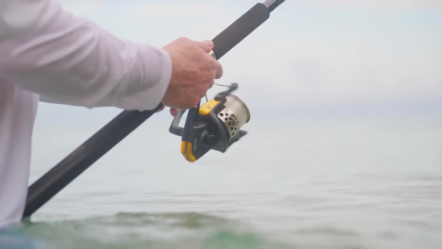 Slow motion close-up of a fisherman reeling in the line on a fishing rod while standing in the ocean