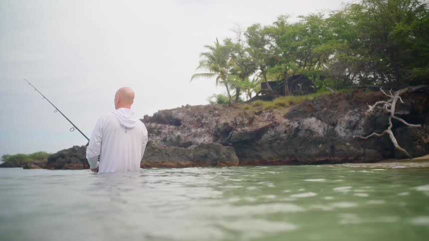 A man stands waist-deep in calm ocean water, holding a fishing rod and looking towards a rocky, tropical shoreline