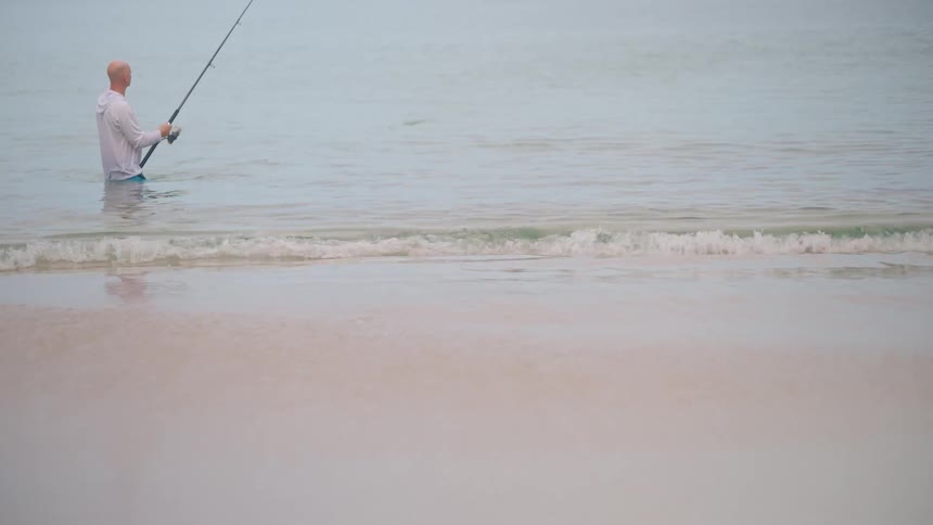 A serene slow-motion shot of a man fishing while standing in the calm ocean as gentle waves lap the sandy shore