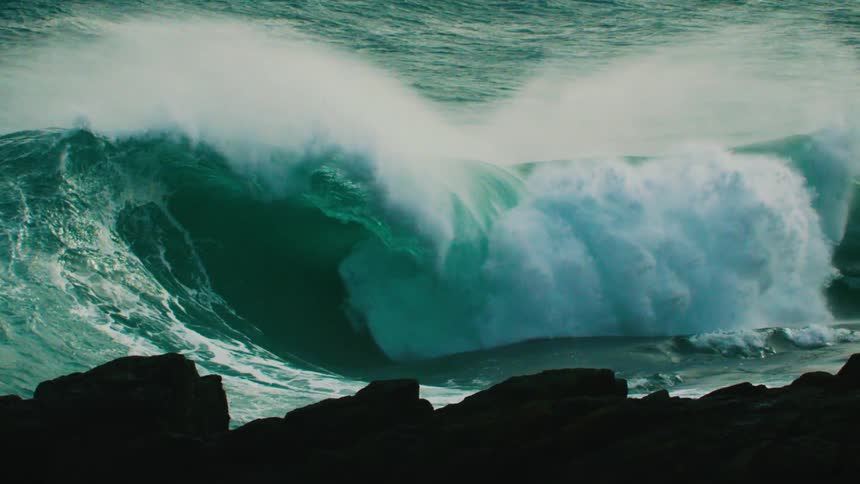 A massive teal ocean wave curls and crashes in dramatic slow motion behind silhouetted rocks