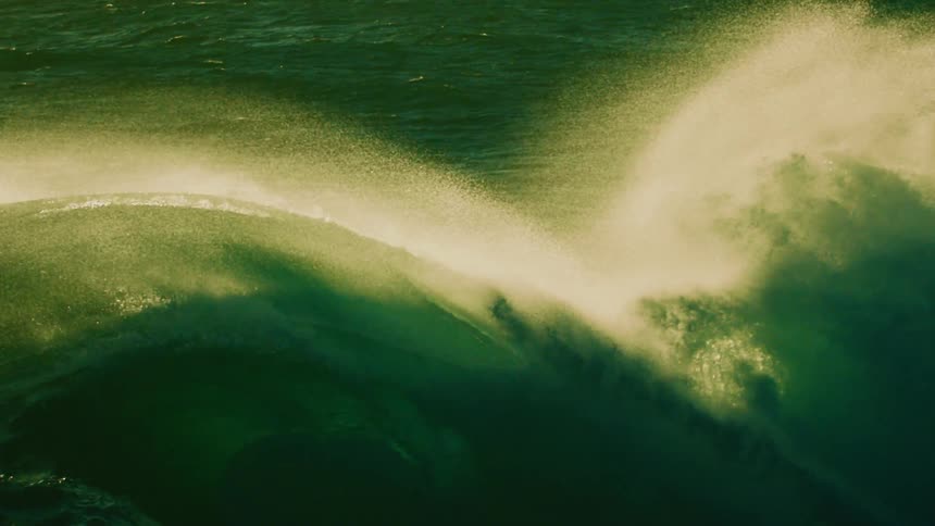 A powerful ocean wave builds and crests in dramatic slow motion, its green water backlit by the sun creating a misty spray