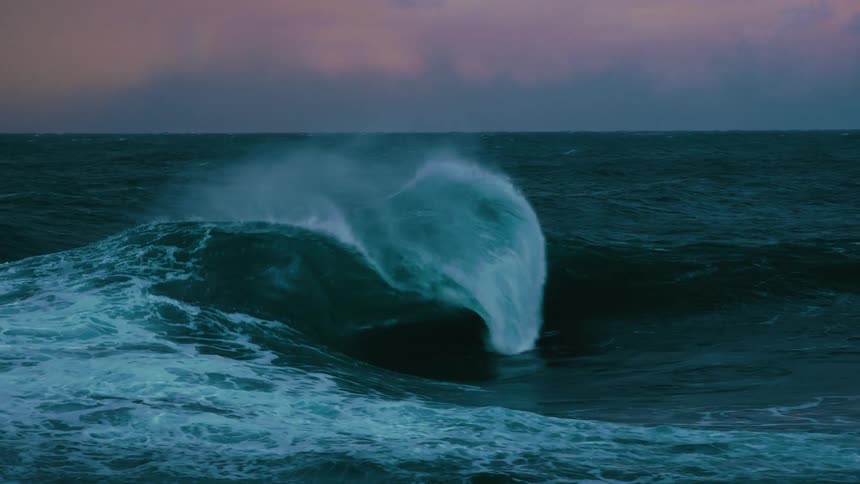 A massive, powerful ocean wave crashes in dramatic slow motion as the wind sprays water from its crest under a moody, purple sky