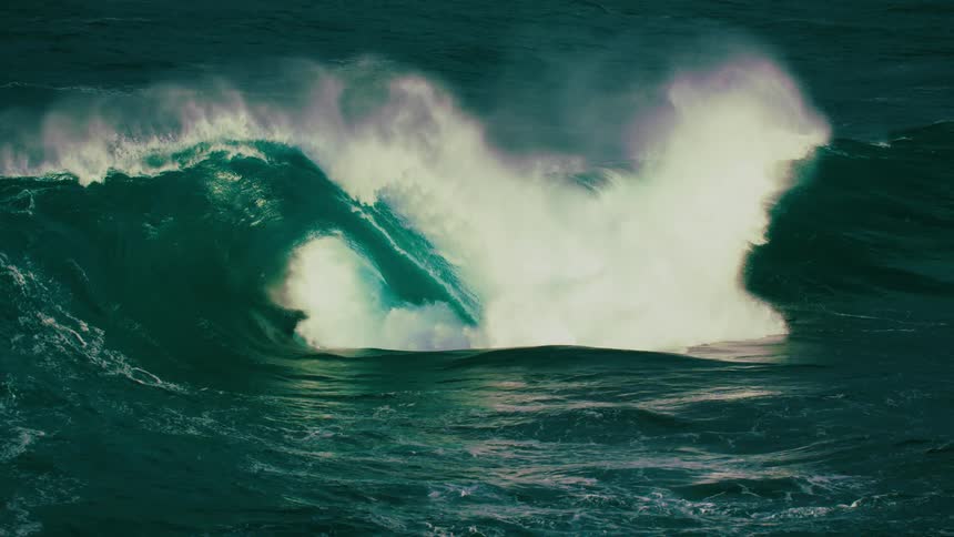 A massive and powerful ocean wave with emerald green water curls and breaks in dramatic slow motion